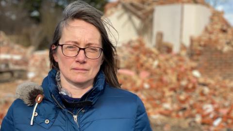 A woman in a blue puffy jacket looks at the camera. She has brown shoulder length hair and is wearing glasses. Behind her is a pile of red brick rubble and a partly demolished house.