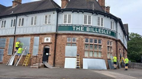 The outside of a two-storey pub is being covered in metal shutters by contractors. The sign outside the pub says 'The Billesley'.