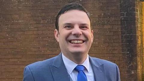 A man, with short brown hair, wearing a grey suit, white shirt and blue tie. He is smiling at the camera. There is a brown brick wall behind him. 