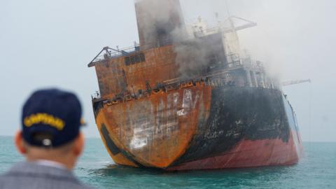 A man looks on as a rusting cargo ship catches fire in Iraqi territorial waters