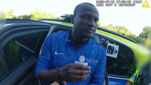 Hadush Kebatu stands in front of police car wearing a blue zipped-up sports jacket. His hands are out in front of him in handcuffs.