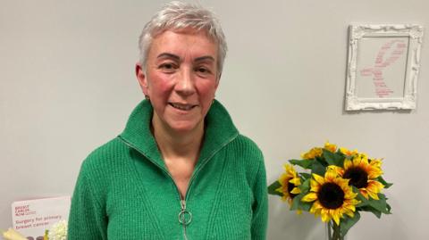 Ann Coffey, who has short white hair and is wearing a green zip-up jumper, smiles at the camera. In the background is a vase with a bunch of sunflowers