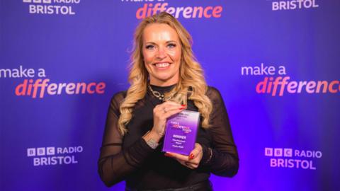 A woman with long, waved, blonde hair holding a purple winner trophy in front of a purple background, which says Make a Difference BBC Radio Bristol on it. She is smiling at the camera and wearing a black, long sleeve, sheer top.