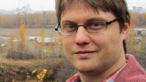 Peter Todd looks at the camera while wearing glasses and a red shirt and standing outside with autumnal trees and a power station in the background