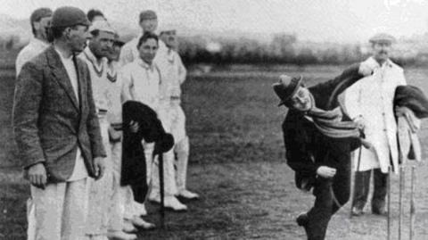A black and white photo of a man wearing a coat, scarf and hat bowling a cricket bowl. A group of men are stood to the left if the photo in cricket whites.