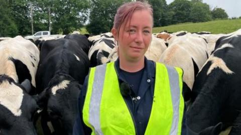 Terri Taylor wearing a high viz vest over her farming overalls is surrounded by black and white cows