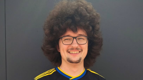 A man with large, fluffy dark curly hair stands smiling against a black wall. 