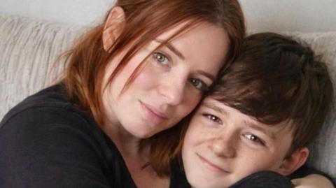 A young teenaged boy with brown hair looks happily into the camera while being embraced by his mother, who has shoulder length reddish brown hair. 
