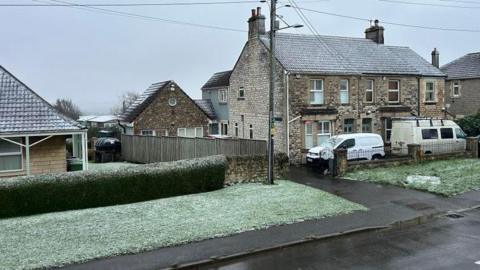 A residential street with a light sprinkling of snow settling on roofs and the grassy verges beside the pavement.