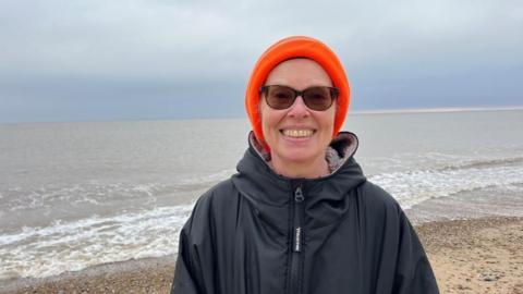 Pam Spychal, dressed in a high visibility orange knitted hat, and a black dry robe. She wears glasses and is pictured on the beach at Great Yarmouth.