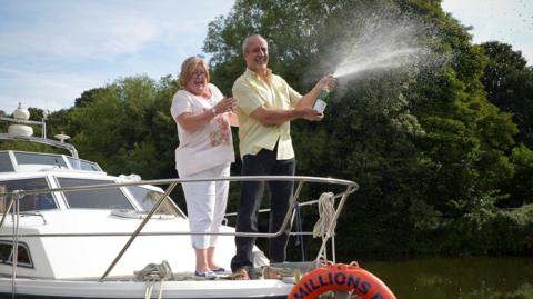 Desiree and Wayne 鶹Լ standing on a boat with an opened bottle of champagne, which is spraying out the top, celebrating their lottery win in August 2014