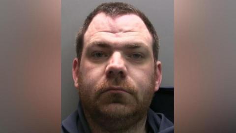 Darrel Carter stares into the camera. He is a ginger coloured beard, short brown hair and has a widow's peak. He is wearing a navy hoodie.