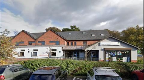 The outside of Church Langley Medical Centre, a brown-bricked building with a partial white stucco front, with three cars parked in front of the glass-panelled door. 