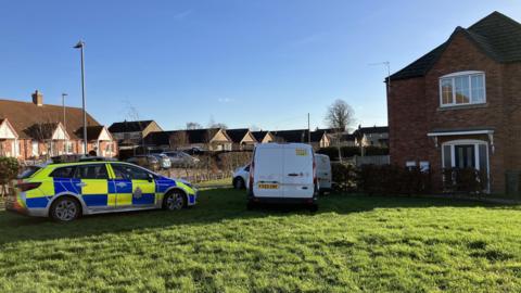 A police car and other vehicles sit on the lawn of a brick house. It is a bright and sunny day.