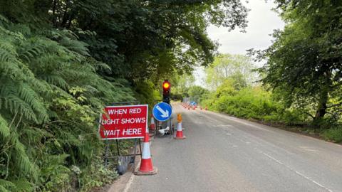 A sign with white letters on a red background reads "when red light shows wait here" on a rural road near Wheddon Cross in Somerset. There are trees and bushes lining each side of the road, which is single lane in each direction. There are also traffic cones and a set of traffic lights visible