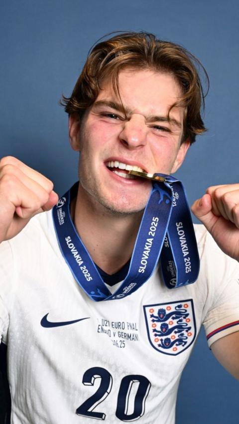 Alex Scott has mid length brown hair in a side parting. He is holding his fists up in a victorious stance and wears a white England football jersey. He has a medal around his neck on a blue ribbon with the metallic medal between his teeth grinning at the camera.