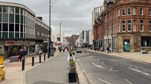 A single carriageway road with plant pots and bollards on the side. Office and retail buildings on both sides of the road. People with shopping bags walk along both sides of the street.