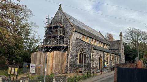 A Victorian church built of square knapped flint, with arched windows with sandstone mullions and a slate roof. The building has been damaged by fire, with plastic sheeting and wooden battens holding temporary repairs in place on the roof. Fencing can be seen around the building, with trees behind.