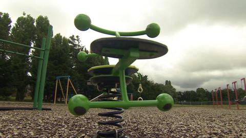 A piece of play park equipment - a spring rider - it is bright green and has two seats. The rest of the park can be seen in the background, with dark clouds overhead. 