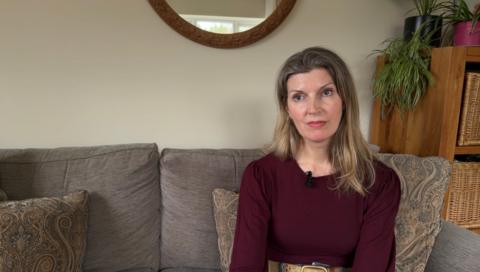 Jenny Tupper, a blonde woman in a burgundy jumper, sits on a grey sofa.