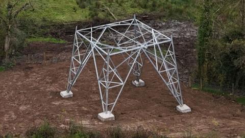 A partially built steel pylon standing in a rural landscape surrounded by uncultivated dark green fields, trees and low hills rolling into the horizon.