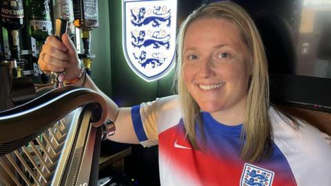 A women smiling and looking directly at the camera. She is wearing a white England football shirt.