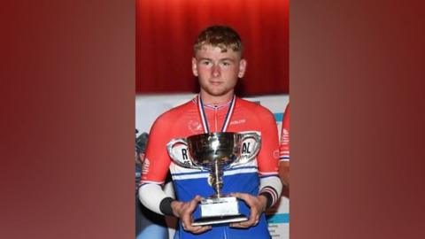 Aidan Worden - a  young man with short, blondish hair and a medal around his neck, holds a trophy. He wears a red, white and blue cycling jersey. 