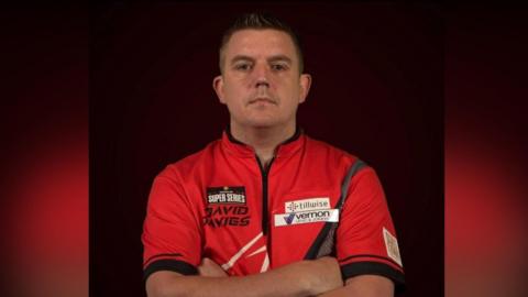 Llun o David Davies yn sefyll gyda'i freichiau wedi croesi o'i flaen wrth edrych at y camera. Mae ganddo wallt brown byr ac mae'n gwisgo crys-t coch