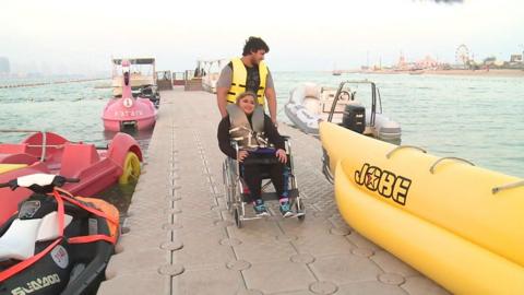 Girl in wheelchair is guided down a pier toward a boat
