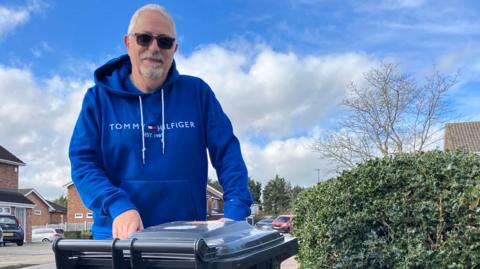 A man with white hair and beard stands behind a black wheelie bin which is out on the pavement. He is wearing a blue hoodie and sunglasses. Next to him on his right is a green hedge and to the left the road is visible with houses and cars parked on the driveways.