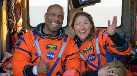 Victor Glover and Christina Koch smile in their orange space suits after returning to Earth. They are sat on a Navy MH-60 Seahawk, the interior of which is visible behind them.