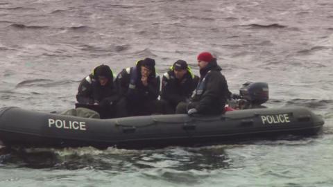 Four men in black coats and life jackets sat on a black police dingy in water.