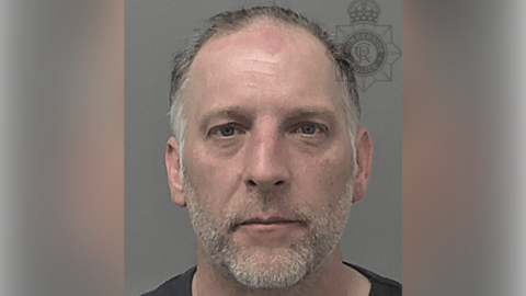 A police custody photo of Nigel Bull. He has short grey hair, a receding hairline and grey facial stubble. He wearing a black t-shirt and is looking at the camera. The wall behind him is grey.