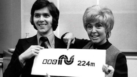 Black and white photo of a man and a woman smiling holding up a sign with "BBC NI 224m" written on it.