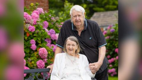 An elderly man and woman are in a garden surrounded by large pink flowers. The man, who has white hair and is wearing a black and blue-trimmed T-shirt, is standing and holding the hand of the woman who is sitting down. She has short fair hair and is wearing a white top and cardigan.