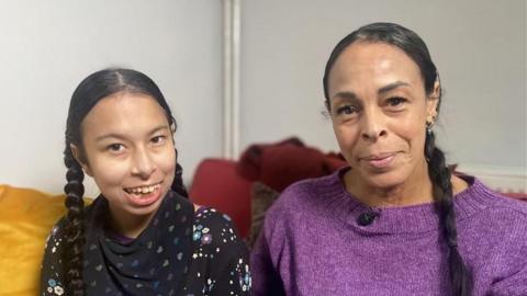 Neave and Natasha are sitting indoors on a sofa. Both have long, dark hair styled in braids/pigtails. Natasha, who is taller, is wearing a purple knitted top with a small microphone clipped near the neckline. Naeve is dressed in a black top patterned with colourful stars and flowers. They are both looking at the camera and smiling.
