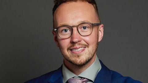 A portrait photo of Adam Dance MP wearing a blue suit, shirt and tie against a dark grey background