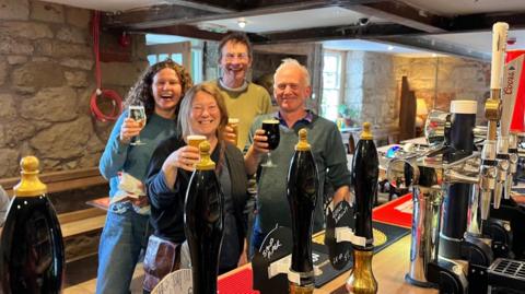 Four people standing in front of a bar in an 18th century pub raising their glasses 