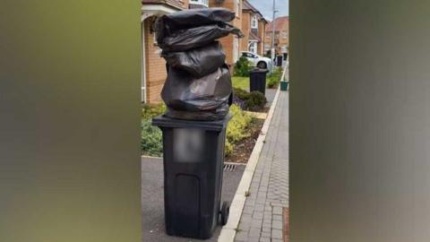 A wheelie bin with bin bags stacked on top of it