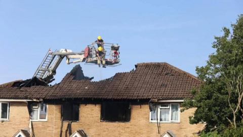 Two firefighters on an areal ladder above a burned roof of a house. It is a clear day.