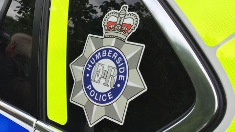 The Humberside Police logo on the back on a police car. It is surrounded by high-visibility yellow and blue stickers. The window has also been tinted and the interior of the car cannot be seen.