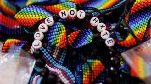 A close-up of handmade bracelets and woven items in rainbow colours, with one beaded bracelet at the centre spelling out “LOVE NOT H8TE” in white letter beads.