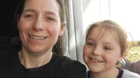 A woman in a black shirt sits next to a small girl in a striped T shirt. They are both smiling at the camera