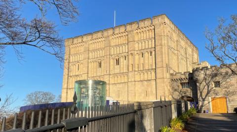 The exterior of Norwich Castle. It is a cube-shaped building made of pale brown stone. A path with railings leads to a smaller, attached part of the castle next to the cube. There is a see-through cylinder structure in front of the castle, containing metal machinery.