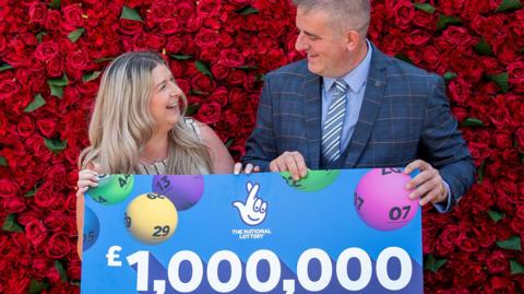 Darren McGuire, who has short greying hair, in a blue check patterned suit with sky blue shirt and blue and white club tie holds a giant cheque for £1m with partner Laura who is wearing khaki sleeveless top