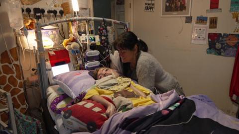 A woman stands over her a man's bed. He is hooked up to ventilators and machines.