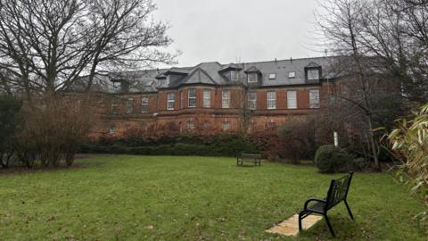 A red brick building with many windows behind a small park with benches, trees and hedges.