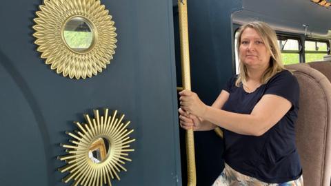 Wendy Salmon looks at the camera and stands at the bottom of the steps on a double decker bus. She is holding on to the handrail, which has been painted gold. The walls of the bus have been painted dark blue and there are gold decorative mirrors on the wall.