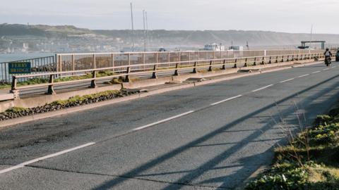 A grey road with white lines and grey fencing with green bushes either side and a number of boats in the background.