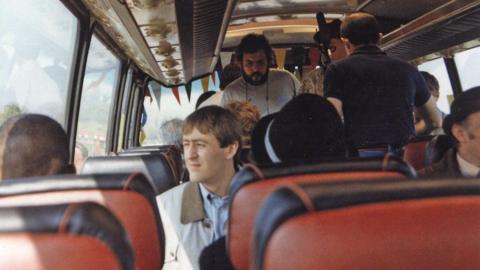 Interior of a coach with rows of red seats, including Rodney Trotter seated by the window among other passengers during a journey.